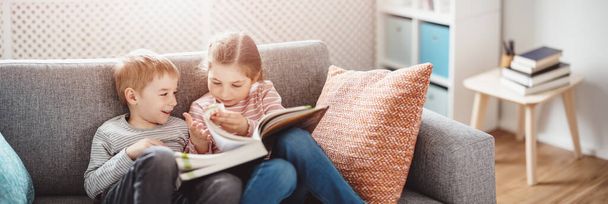 Cute children sitting on the soft sofa and reading books - Zdjęcie, obraz