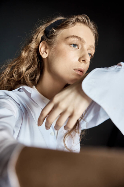 portrait shoot in the studio on a dark background of a beautiful young blonde girl with big blue eyes, long wavy hair, she is dressed in a white cotton shirt with a T-shirt under it and posing - Zdjęcie, obraz