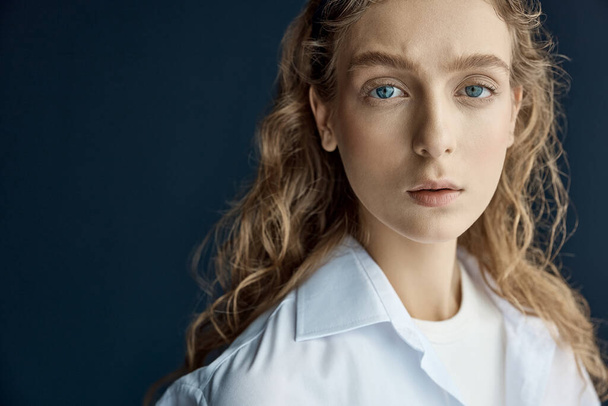 portrait shoot in the studio on a dark background of a beautiful young blonde girl with big blue eyes, long wavy hair, she is dressed in a white cotton shirt with a T-shirt under it  - Φωτογραφία, εικόνα