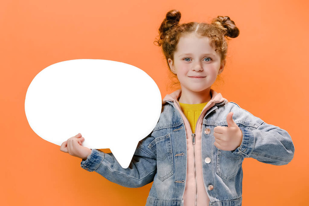 Funny nice little girl kid dressed in denim jacket hold white empty blank Say cloud, speech bubble showing thumb up, isolated on pastel orange color background with copy space. People emotion concept - Foto, imagen