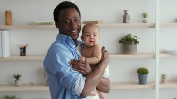 陽気なアフリカ系アメリカ人の父親を保持し、彼の愛らしい新生児の赤ちゃんをlullie 、リビングルームのインテリアでカメラで笑顔 - 映像、動画