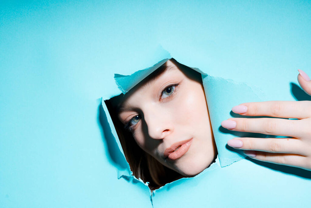 Beautiful face of a girl with makeup peeking out of a hole in a blue background revealing her hand - Fotoğraf, Görsel