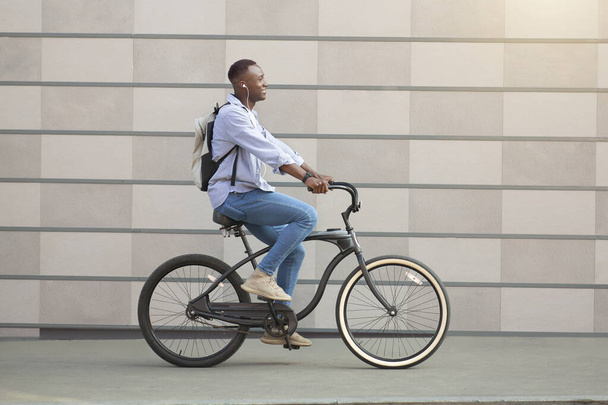 Seitenansicht eines coolen Schwarzen mit Kopfhörern, der auf seinem Fahrrad in der Nähe der Backsteinmauer in der Innenstadt rast - Foto, Bild