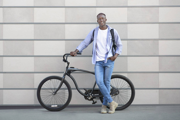 Retrato de cuerpo entero del negro feliz con la bicicleta moderna de pie en la calle de la ciudad y mirando a la cámara - Foto, Imagen