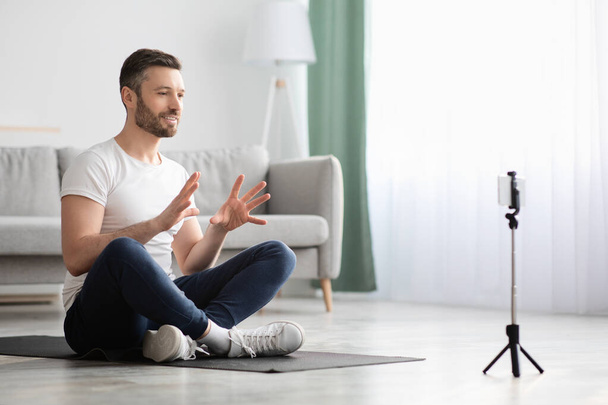 Man fitness coach sitting on yoga mat, broadcasting with smartphone - Photo, image