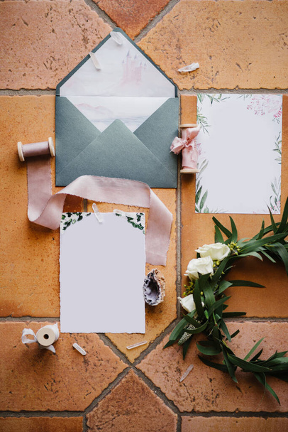 Op een bruine tegel - een open grijze envelop, blanco ansichtkaarten met ornamenten, strengen linten, een schelp en een krans van twijgen met een roos - Foto, afbeelding