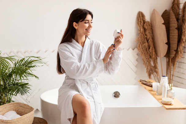 Vrouw Holding Cream hydraterende huid zitten op bad in de badkamer - Foto, afbeelding