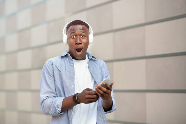 Impresionado hombre afroamericano con dispositivo móvil y auriculares mirando a la cámara cerca de la pared de ladrillo, espacio libre - Foto, imagen