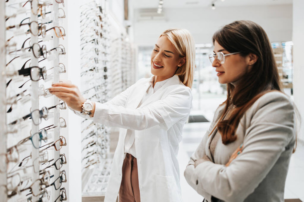 Beautiful and fashionable woman choosing eyeglasses frame in modern optical store. Female seller specialist helps her to make right decision. - Fotoğraf, Görsel