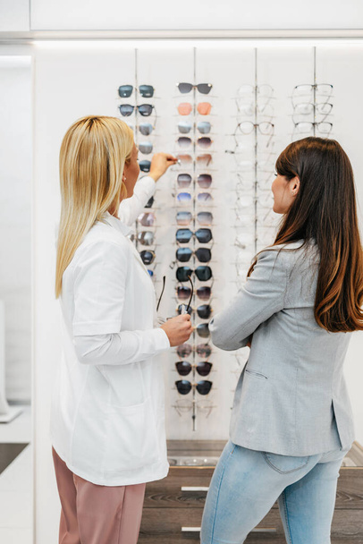 Beautiful and fashionable woman choosing eyeglasses frame in modern optical store. Female seller specialist helps her to make right decision. - 写真・画像