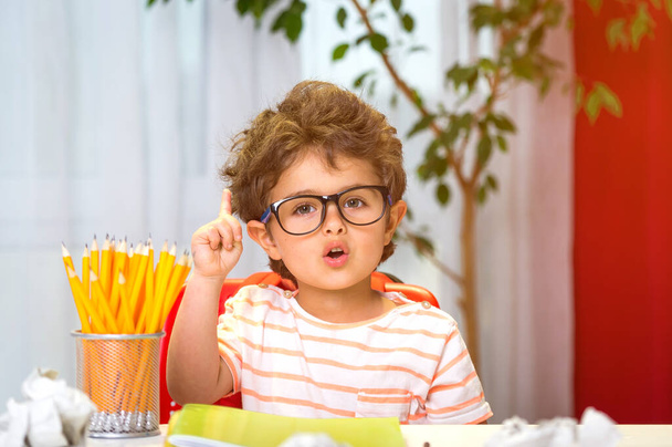 Okula dönelim. Gözlüklü mutlu, sevimli, çalışkan çocuk kapalı bir masada oturuyor. Akıllı çocuk sınıfta parmak kaldırarak öğreniyor. İnek öğrenci. - Fotoğraf, Görsel