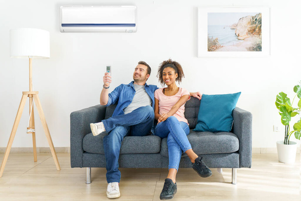 Fröhliches Paar sitzt auf Sofa und schaltet Klimaanlage ein - Foto, Bild