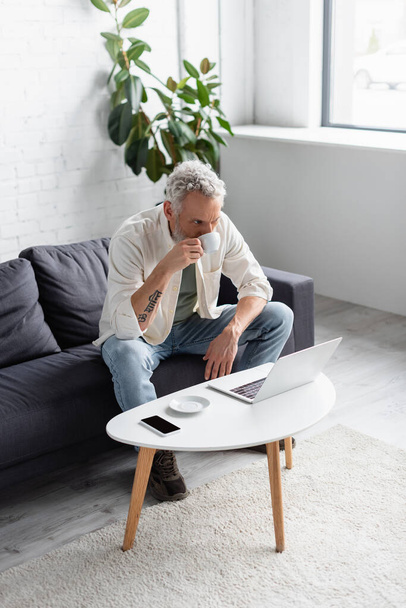 uomo barbuto bere caffè e utilizzando il computer portatile mentre seduto sul divano vicino smartphone con schermo bianco sul tavolino - Foto, immagini
