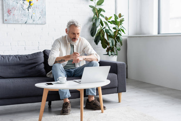 bearded man pointing at laptop while sitting on couch near cup of coffee and smartphone with blank screen on coffee table - 写真・画像