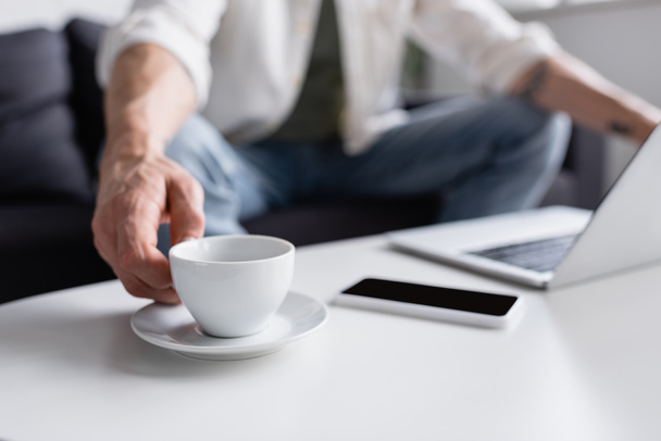 vista cortada do homem atingindo xícara de mesa de café perto de gadgets  - Foto, Imagem
