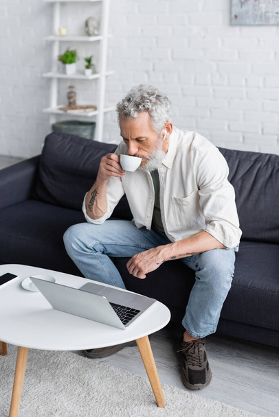 bearded man in wireless earphones drinking coffee and looking at laptop  - Zdjęcie, obraz