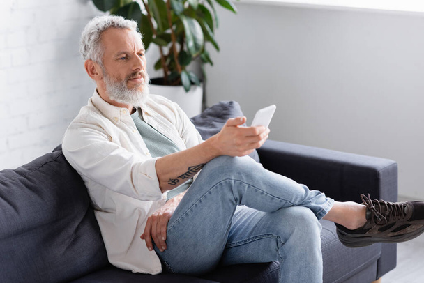 bearded man messaging on smartphone while sitting on couch - Photo, image