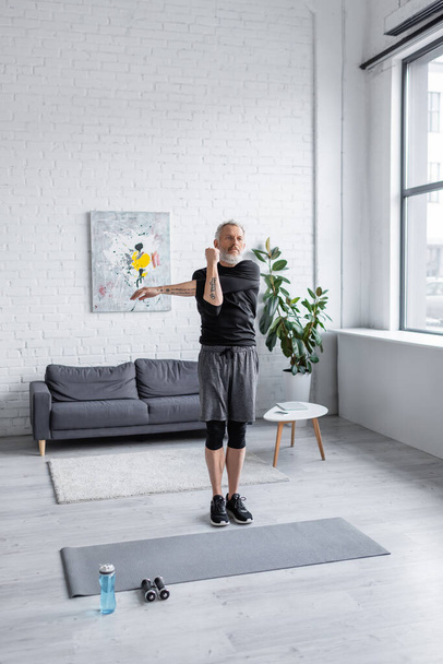 teljes hossza szakállas férfi gyakorló fitness mat közelében súlyzók a nappaliban  - Fotó, kép