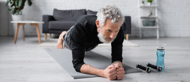 bärtiger Mann beim Planken auf Fitnessmatte in der Nähe von Hanteln im Wohnzimmer, Banner - Foto, Bild