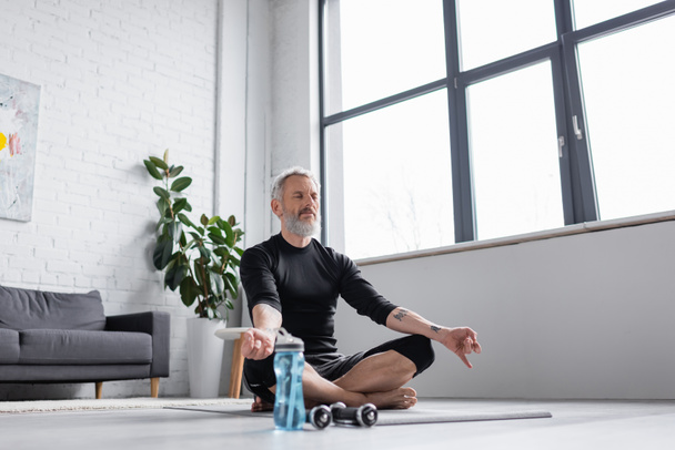 brodaty mężczyzna z siwymi włosami medytujący na macie do jogi przy hantlach w salonie - Zdjęcie, obraz