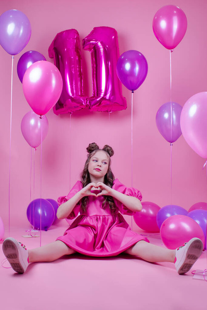 słodka brunetka dziewczyna z różowym makijażu i w różowej skórzanej sukience. blisko kolorowych balonów w kształcie jedenastki. na różowym tle.koncepcja radości, przyjęcie, urodziny - Zdjęcie, obraz