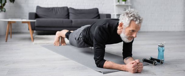 uomo scalzo con i capelli grigi che fa tavola sul tappeto fitness vicino ai manubri in soggiorno, banner - Foto, immagini