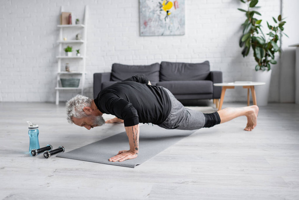 Barfuß-Mann mit grauen Haaren trainiert auf Fitnessmatte neben Hanteln im Wohnzimmer - Foto, Bild