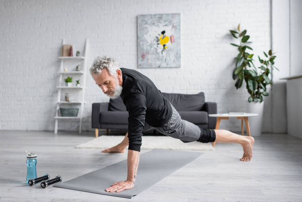 Sportlicher Mann mit grauen Haaren trainiert auf Fitnessmatte neben Hanteln im Wohnzimmer - Foto, Bild