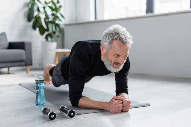 strong man with grey hair doing plank on fitness mat near dumbbells and sports bottle in living room - Photo, Image