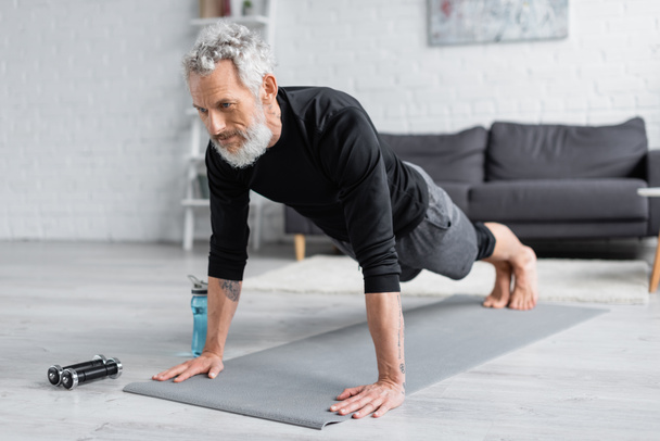 tattooed man with grey hair doing plank on fitness mat near dumbbells and sports bottle in living room - Photo, Image