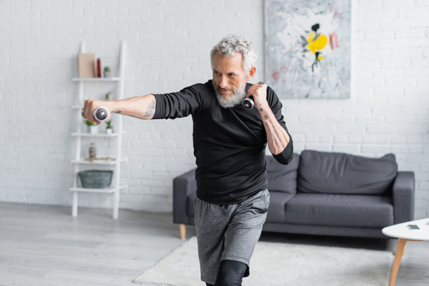 tattooed and bearded man working out with dumbbells in living room - Valokuva, kuva