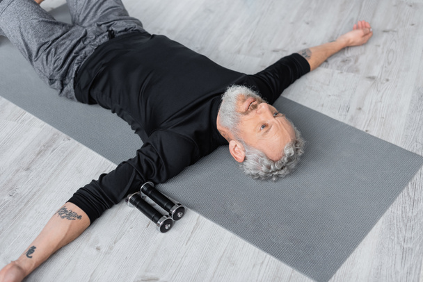 hoge hoek uitzicht van bebaarde man liggend op fitness mat in de buurt halters  - Foto, afbeelding