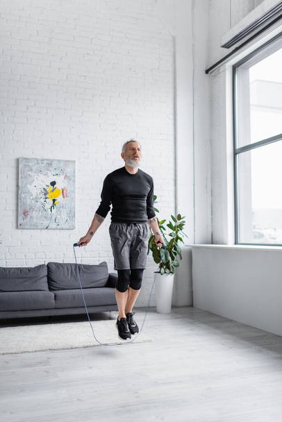vousatý muž s šedými vlasy pracuje s skákacím lanem v obývacím pokoji  - Fotografie, Obrázek