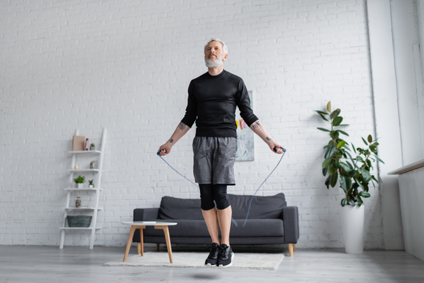 bearded man in sportswear exercising with jumping rope in modern living room  - Φωτογραφία, εικόνα