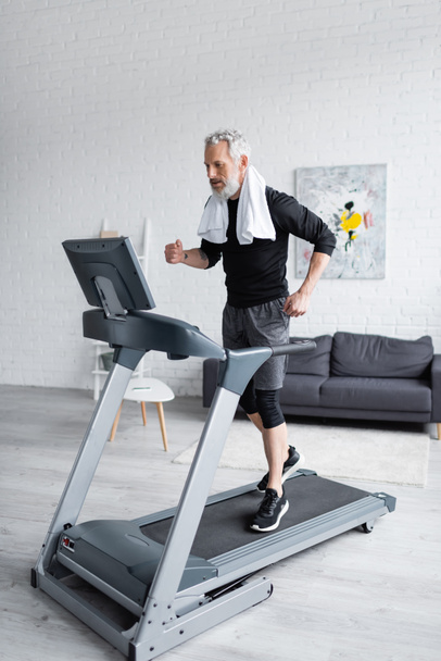 full length of bearded man with white towel jogging on treadmill at home  - Fotoğraf, Görsel