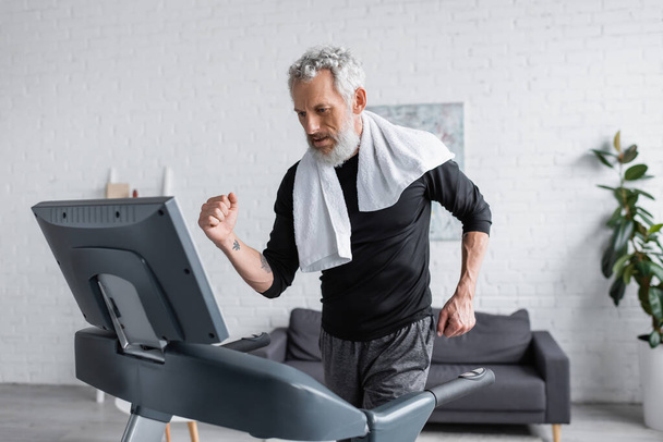 bearded man with white towel running on treadmill at home  - Foto, afbeelding