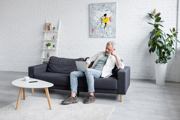 Mann mit grauen Haaren benutzt Laptop, während er auf Couch neben Tasse Kaffee und Smartphone mit leerem Bildschirm auf Couchtisch sitzt - Foto, Bild