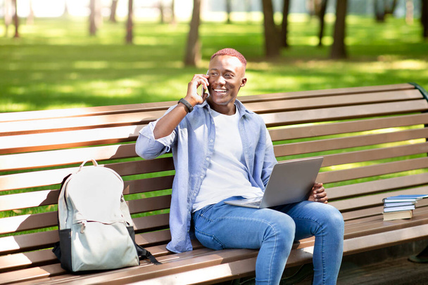 Концепция электронного обучения. Чернокожий студент с ноутбуком разговаривает по смартфону на скамейке в парке - Фото, изображение
