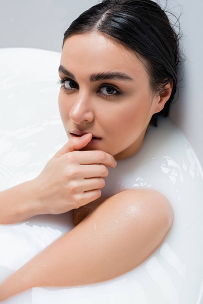 mujer seductora mirando a la cámara y tocando el labio mientras toma un baño con leche - Foto, Imagen