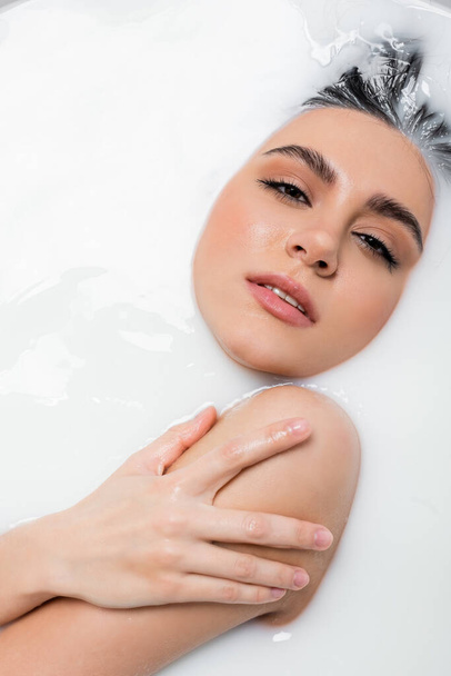 sensual woman touching shoulder and looking at camera while relaxing in milk bath - Photo, Image