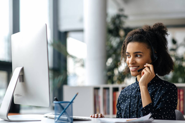 Confident beautiful african american girl in headset, manager, call center worker ή freelancer, κάθεται στο τραπέζι, χρησιμοποιώντας υπολογιστή, επικοινωνώντας με συνάδελφο ή πελάτη μέσω video link, χαμογελώντας - Φωτογραφία, εικόνα