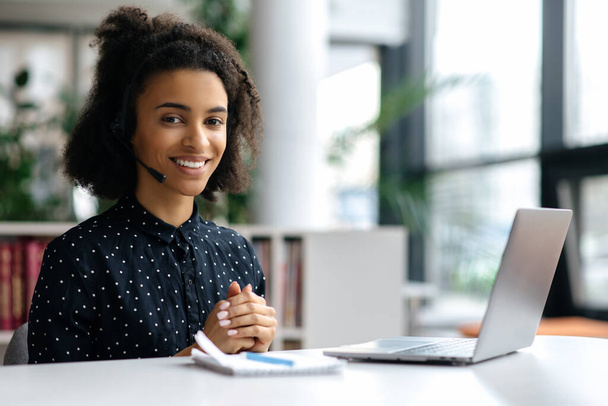 Ritratto di giovane donna afroamericana attraente in auricolare, manager, call center lavoratore o freelance, seduta in ufficio, vestita con stile, usando il computer portatile, guardando la macchina fotografica, sorridendo - Foto, immagini