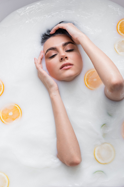 bovenaanzicht van vrouw met gesloten ogen ontspannen in melkachtig bad met gesneden citrusvruchten - Foto, afbeelding