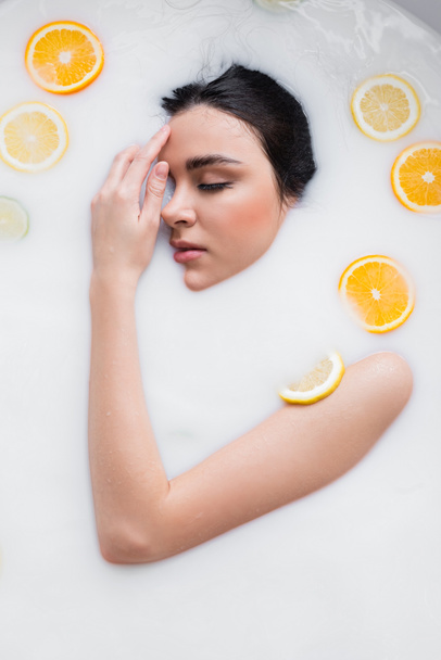 young woman holding hand near face while relaxing in milk bath with sliced lemons and oranges - Photo, Image