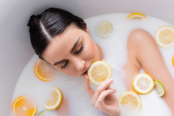overhead view of woman holding lemon slice while relaxing in milk bath - Photo, Image