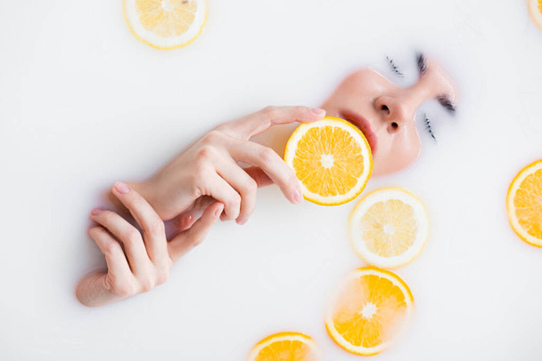 vista superior de la mujer sosteniendo rebanada de naranja en baño de leche - Foto, Imagen