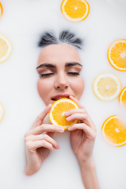 young woman holding slice of fresh orange while relaxing in milk bath - Photo, Image