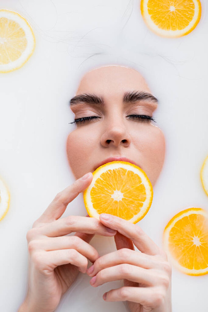 Süt banyosu yaparken elinde portakal dilimi tutan gözleri kapalı bir kadın. - Fotoğraf, Görsel