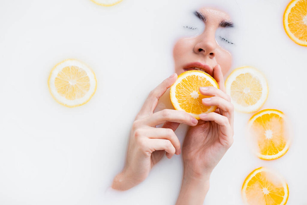 high angle view of woman holding orange slice while immersing face in milk bath - Foto, Imagen