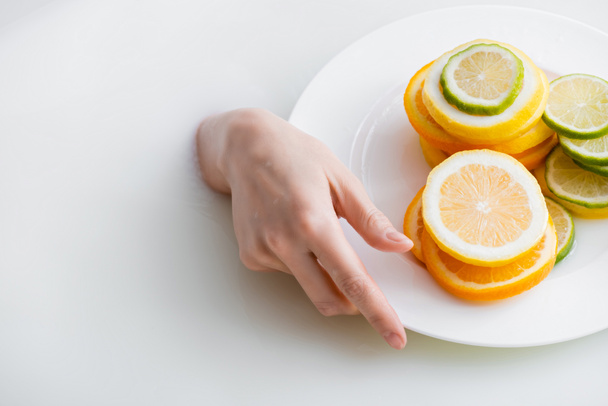 vista parcial de la mano femenina en baño de leche cerca de la placa con limón en rodajas, lima y naranja - Foto, Imagen